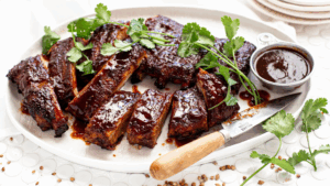 Oven baked pork ribs coated in glossy barbecue sauce, sliced on a serving platter with fresh coriander garnish and dipping sauce.